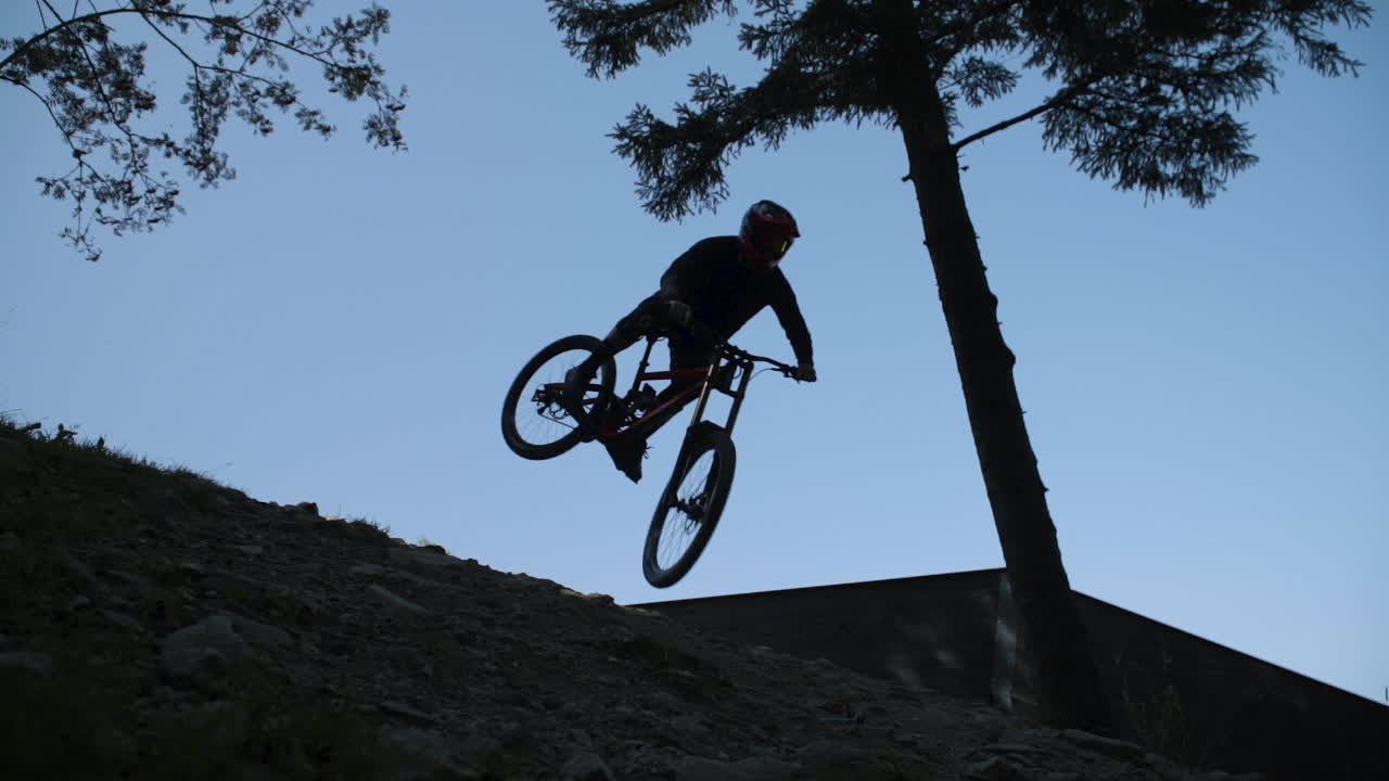 Slow motion shot of downhill racer hitting a big jump with a whip, only silhouette is visible against blue sky. Downhill park in Semmering Austria. Footage slowed down eight times