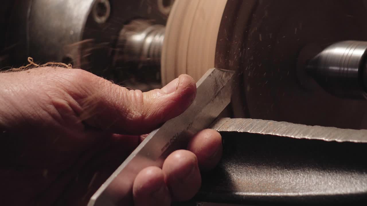 Close-up of a woodturner using a gouge chisel to shape spinning wood on a lathe. Artisan woodworking process creating shavings in a workshop setting