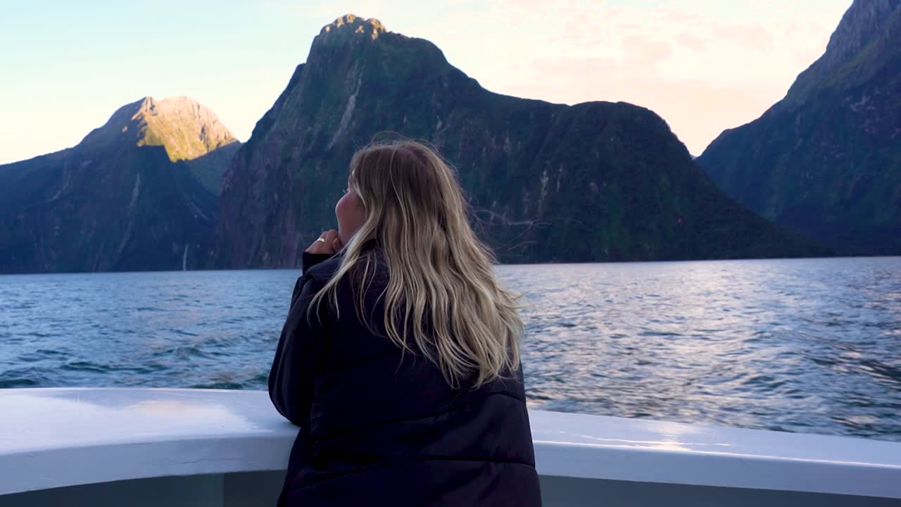 A woman stands at the front of a boat sailing through Milford Sound in Fiordland National Park, New Zealand. She is taking in the towering cliffs and waterfalls all around the fjord