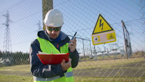 Ingeniero-Masculino-Hablando-Por-El-Walkie-Talkie-Y-Leyendo-En-El-Portapapeles-En-La-Subestación-Eléctrica,-Dinámico-Portátil