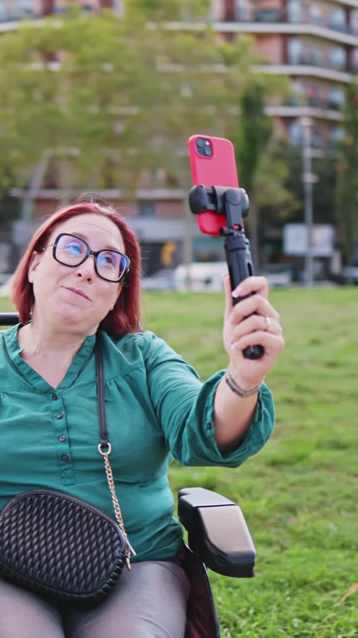 Donna felice in sedia a rotelle che fa vlogging con lo smartphone al parco.