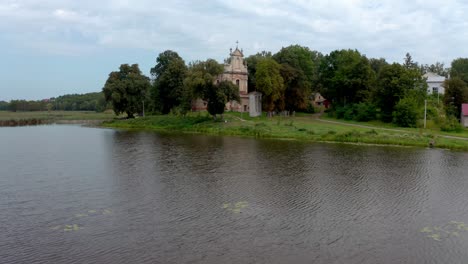 Hodovytsya,-Ukraine---Katholische-Kirche-Aller-Heiligen,-Umgeben-Von-Grünen-Bäumen,-Errichtet-In-Der-Nähe-Des-Ruhigen-Sees-Unter-Dem-Bewölkten-Himmel---Weitwinkelaufnahme