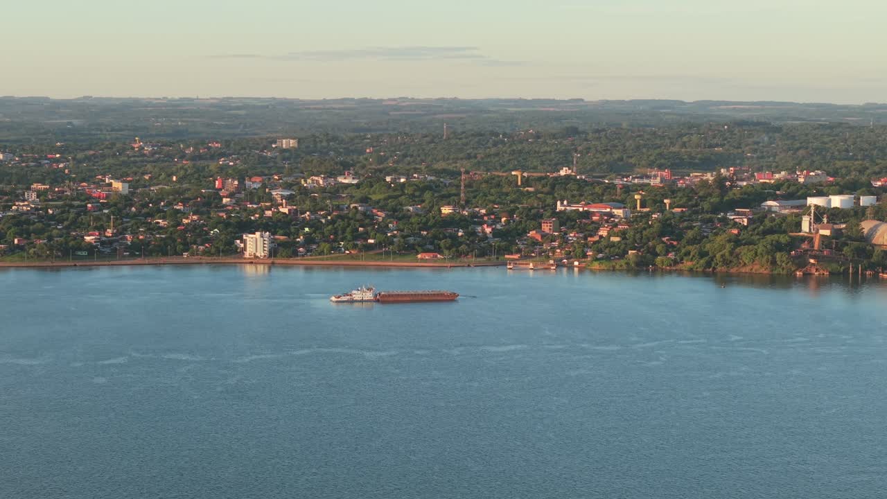 Aerial view from Posadas shows anchored grain barge on Paraná River facing Encarnación, Paraguay, with urban skyline, red roofs, trees and distant hills under soft morning light