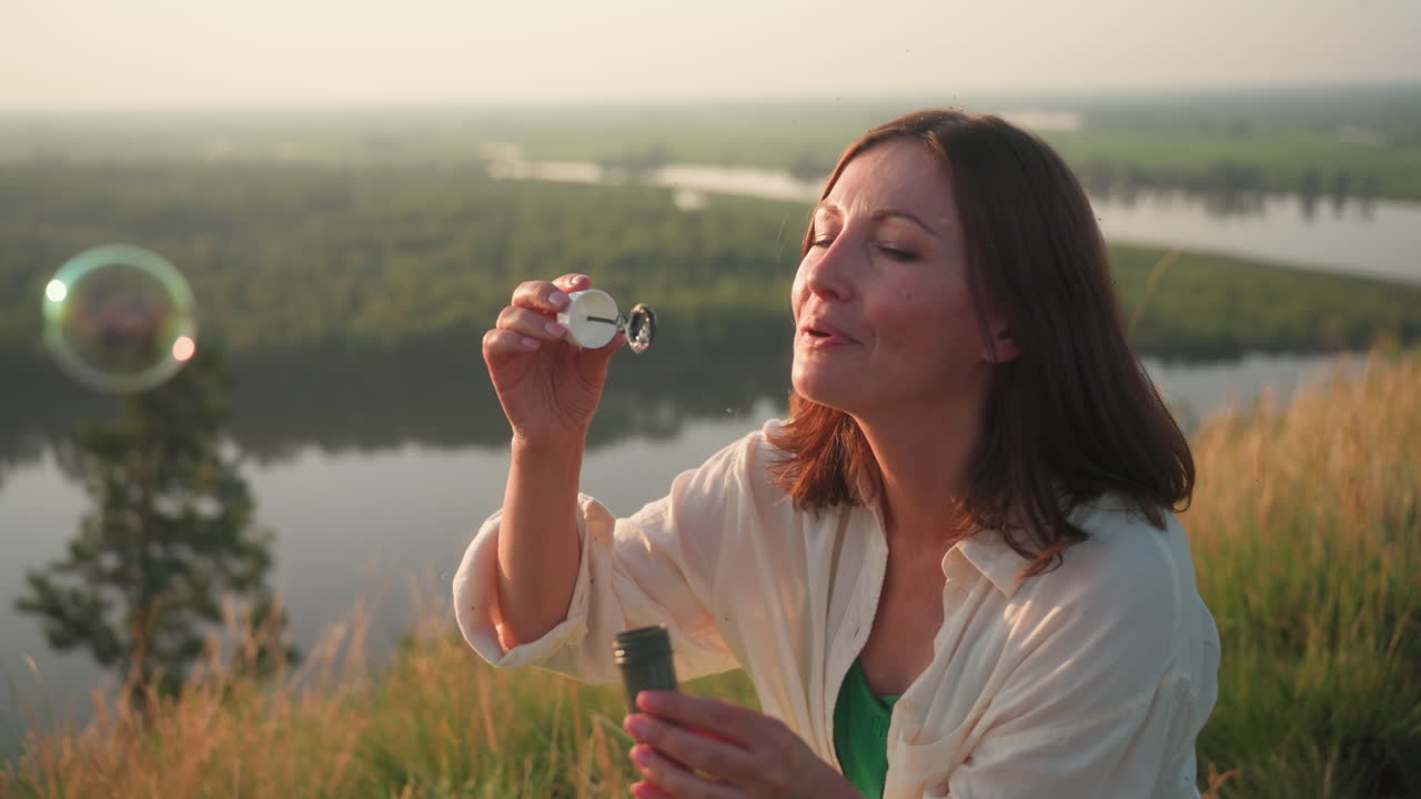 Young woman blowing soapy bubbles while seated on grassy hillside overlooking winding river valley at sunset, holding bubble container in hand, surrounded by tall grasses swaying in gentle breeze