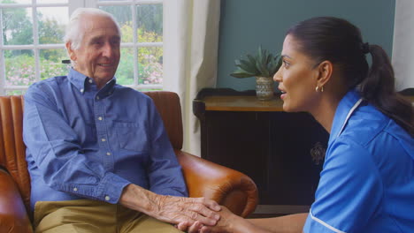 female care worker in uniform holding hands of senior man sitting in care home lounge