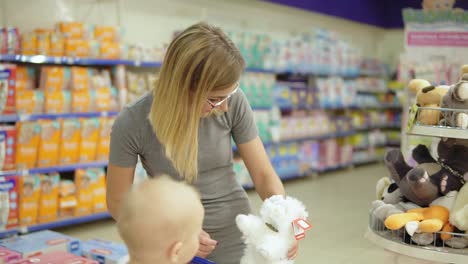 Lächelnde-Mutter-Und-Ihr-Kind-In-Der-Spielzeugabteilung-Im-Supermarkt,-Die-Spielzeug-Auswählen.-Süßes-Kleines-Baby,-Das-In-Einem-Einkaufswagen-Sitzt-Und