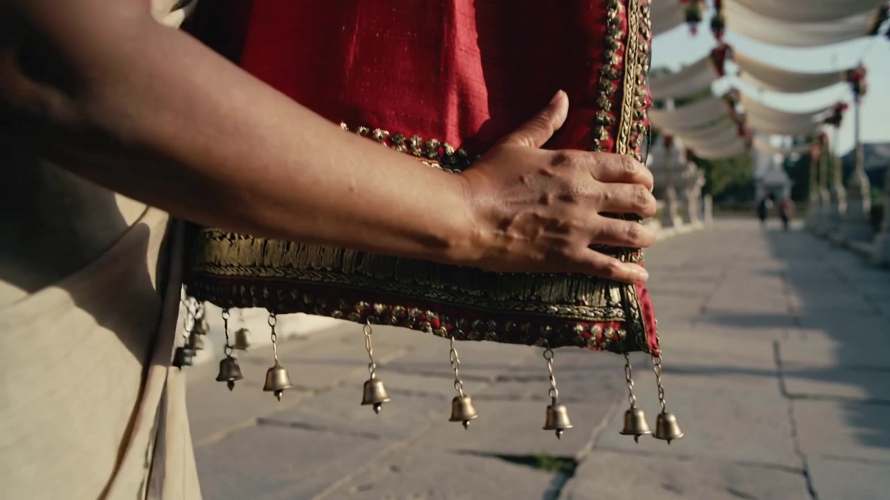Donna in sari beige che cammina, sistemando una pochette rossa ricamata con campanelle pendenti su un sentiero di pietra.