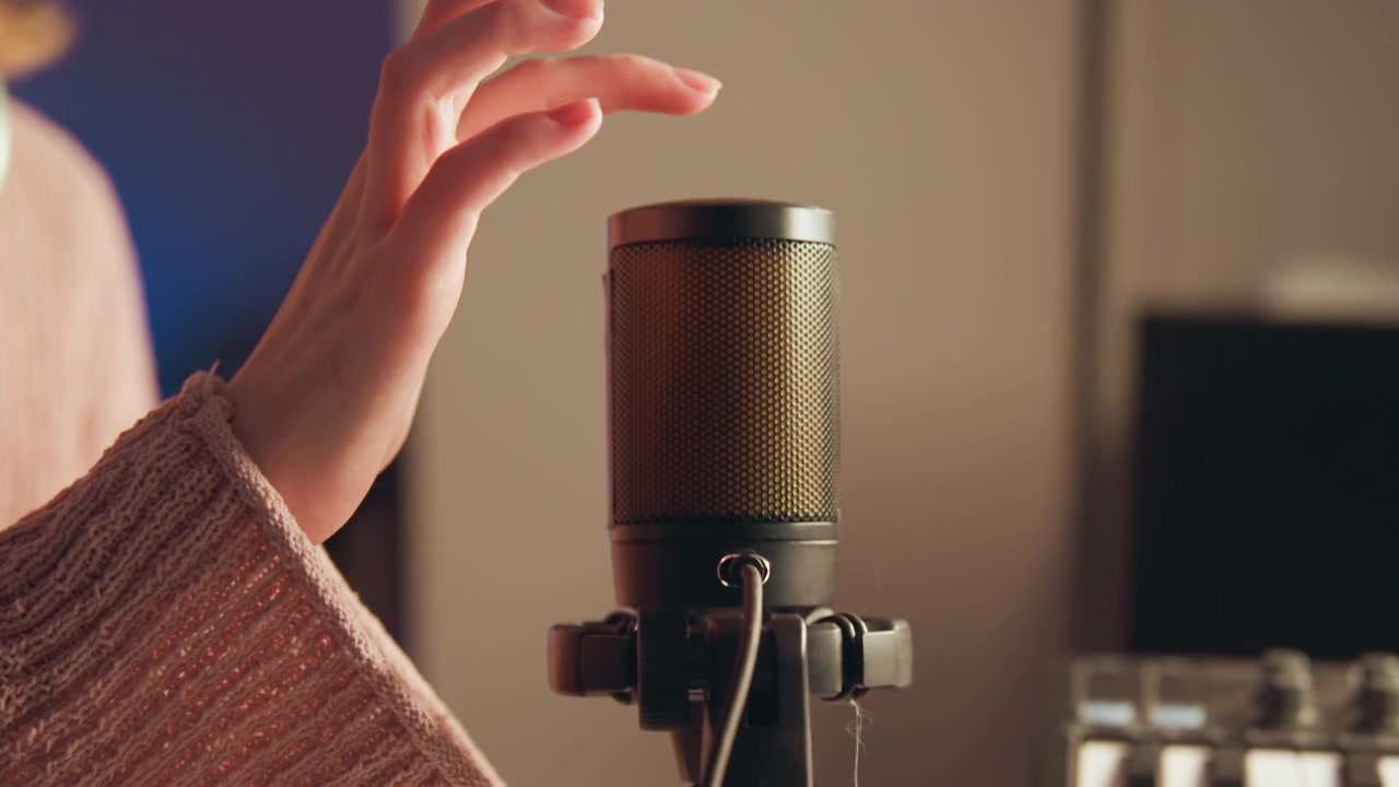 Un microfono a condensatore verticale si trova in uno studio casalingo caldo e illuminato delicatamente, mentre una mano si muove vicino alla griglia durante la configurazione, suggerendo una registrazione, un doppiaggio, un podcast o una conversazione in streaming.
