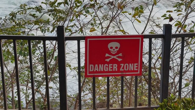 Close view of red danger zone warning sign with skull symbol mounted on metal railing, river water visible in background creating a strong safety alert scene outdoors