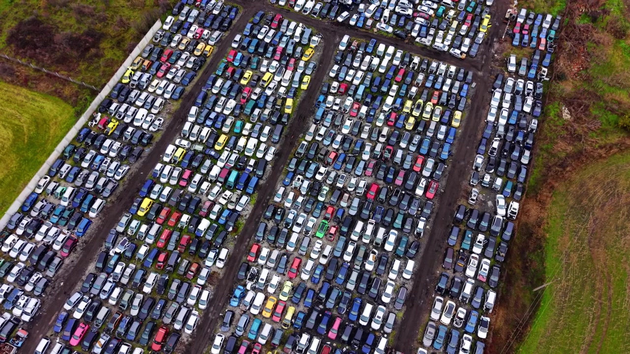 Desguace de coches a gran escala visto desde arriba. Carriles diagonales de vehículos destrozados se extienden hacia campos verdes bajo un cielo invernal nublado.