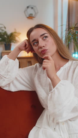 thoughtful young woman in dress touching chin thinking about where to go for weekend sitting on sofa