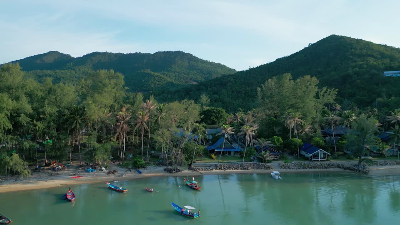 Aerial pullback over lush green hills and coastline with scattered resorts, calm waters reflecting the vibrant landscape as boats float in sea