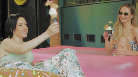 two young women enjoy ice cream in a relaxed atmosphere next to an inflatable pool in front of a contemporary ice cream parlor.
