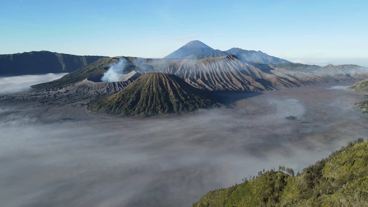 Aerial drone footage of Mount Bromo volcano with smoke rising, surrounded by misty sea of sand and mountain range under clear blue sky