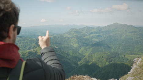 a close up camera shot from behind of a man with sunglasses looking from the top of the mountain into the valley and pointing at the other mountains to identify them