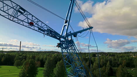 rear strut and front strut of a blue luffing construction crane against a blue sky and clouds with forest in background