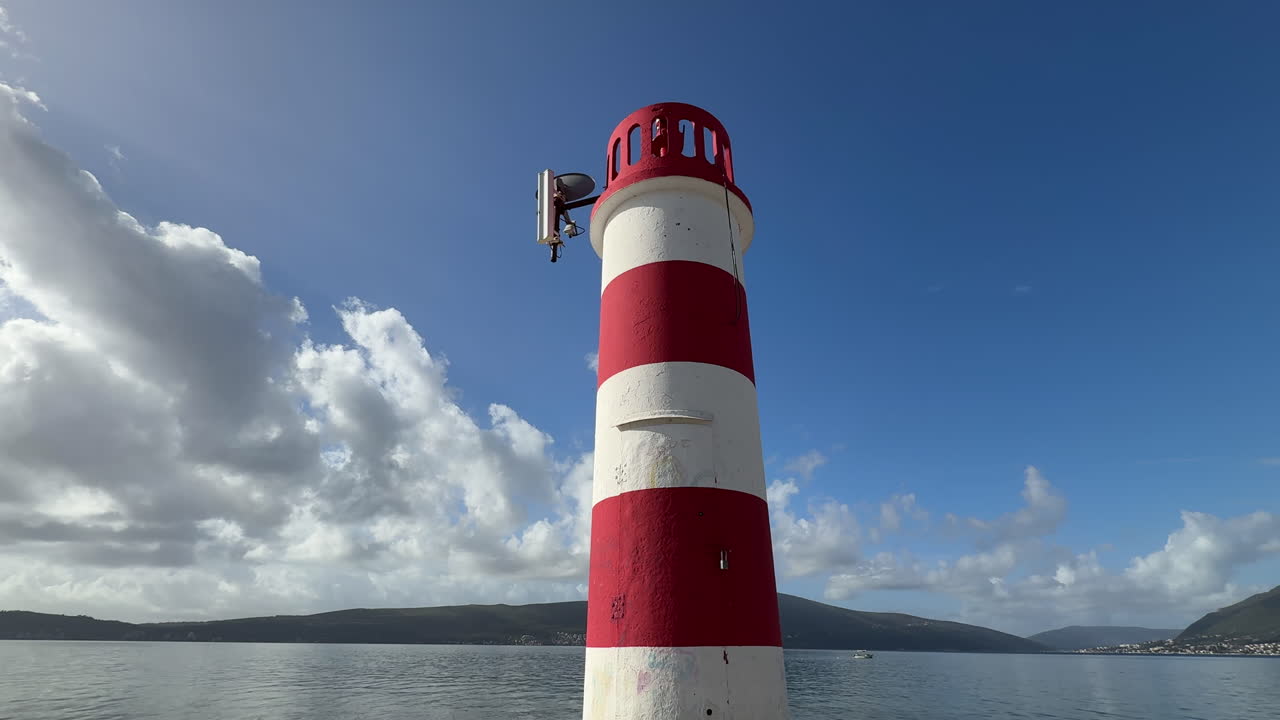 A small red-white coastal beacon stands by the sea on a clear sunny day, overlooking calm blue water, distant hills, and a peaceful shoreline. Serene maritime scenery ideal for travel and nature