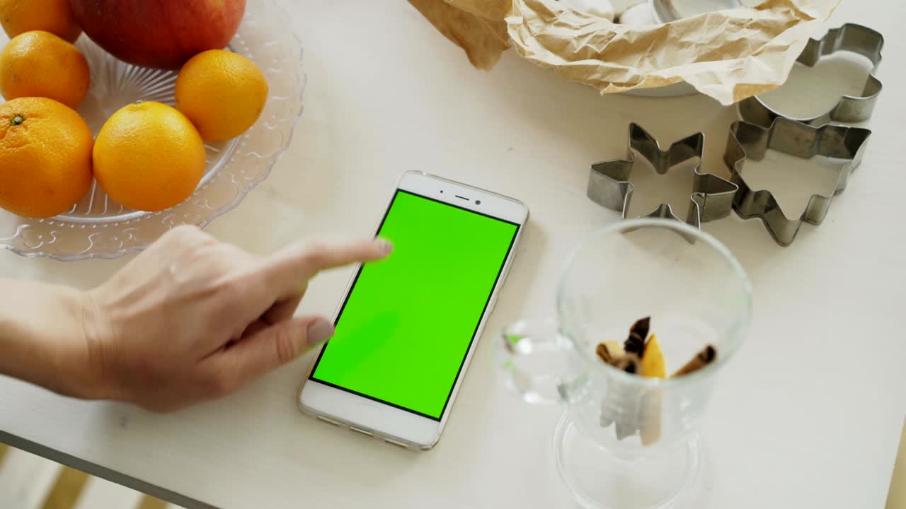 primer plano de la mano de la mujer navegando por el teléfono inteligente con pantalla verde en la mesa de la cocina en casa