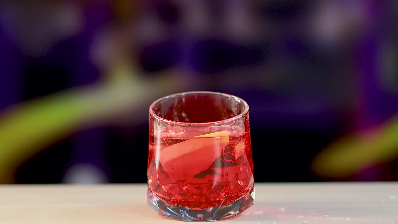 Un cocktail Negroni è guarnito con una scorza d'arancia su un bancone da bar sotto un'illuminazione vivace.