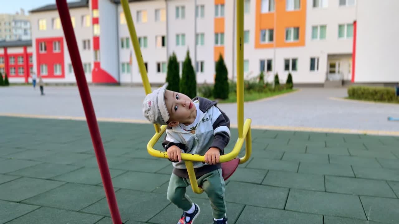 Sweet Caucasian baby swinging on the playground. Cute boy bending head looking at camera. Blurred buildings at backdrop.