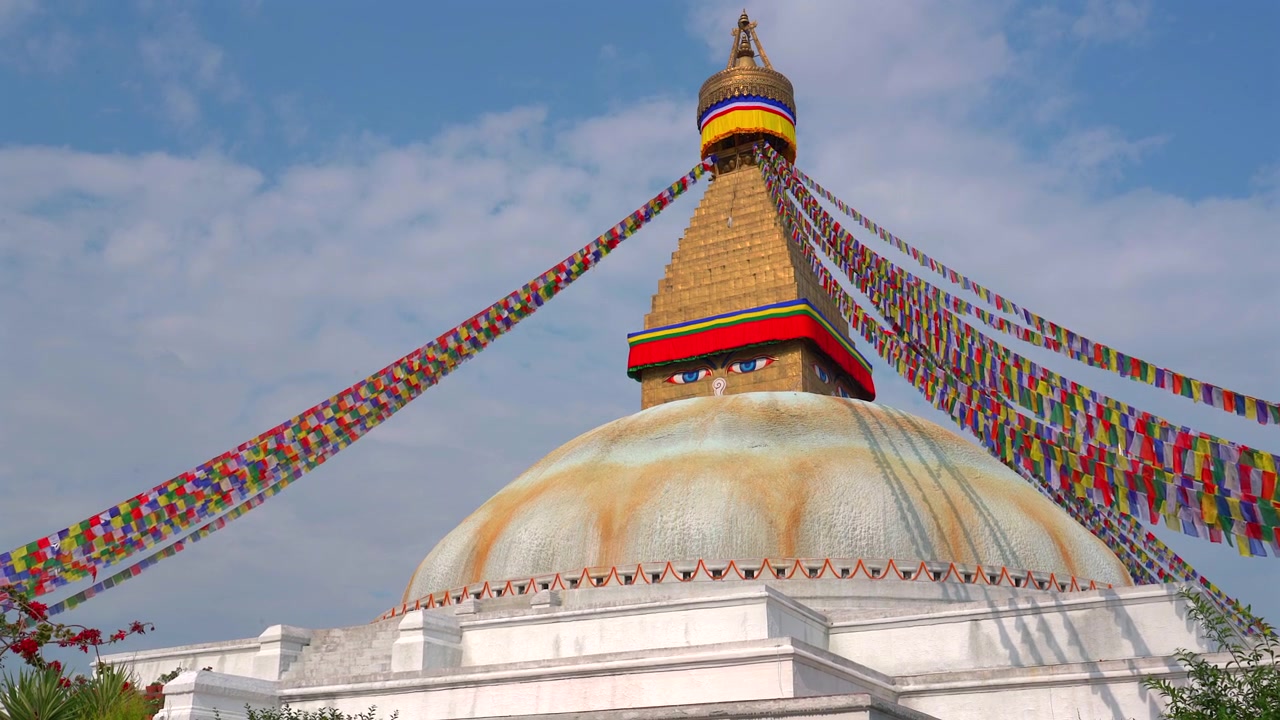 Seurantakuva Boudhanathin stupan suuresta valkoisesta kupolista ja ikonisesta kultaisesta tornista, jotka ovat koristeltu eloisilla rukouslipuilla sinisen taivaan alla Kathmandussa, Nepalissa.