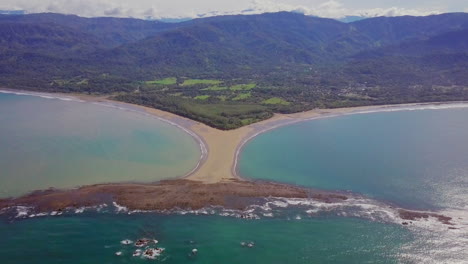 aerial shot pulling away from the whale tail shaped rocky point of punta uvita surrounded by bright blue ocean waves and lush rainforest in the distance in south puntarenas, costa rica