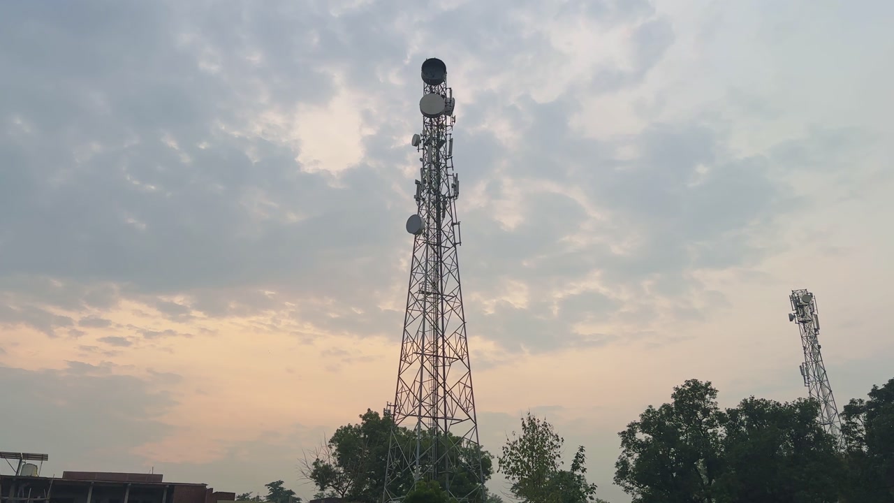 A shot begins with surrounding greenery and buildings, then tilts up to reveal tall telecom towers and antennas silhouetted against a cloudy evening sky.