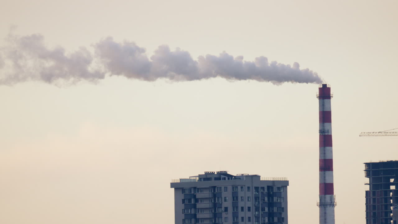 Skyline urbano con edifici residenziali e una ciminiera industriale che emette fumo al tramonto.