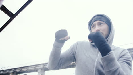 vista inferior del hombre caucásico con capucha gris golpeando un saco de boxeo al aire libre en una fábrica abandonada en una mañana nublada