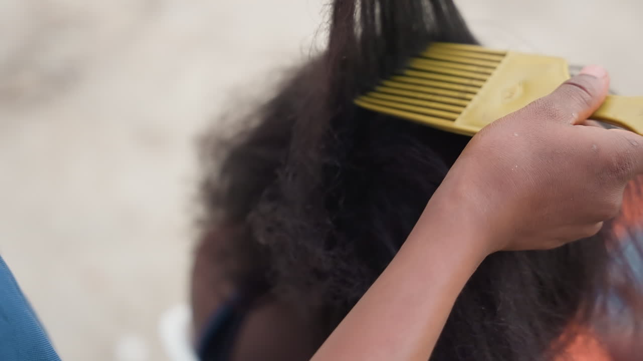 Gros plan sur des mains peignant des cheveux naturels, un soignant démêlant délicatement le cuir chevelu texturé d'un enfant avec un peigne à dents larges dans un cadre intérieur calme, douce lumière du soleil, mouvements patients, routine hydratante, bienveillance.