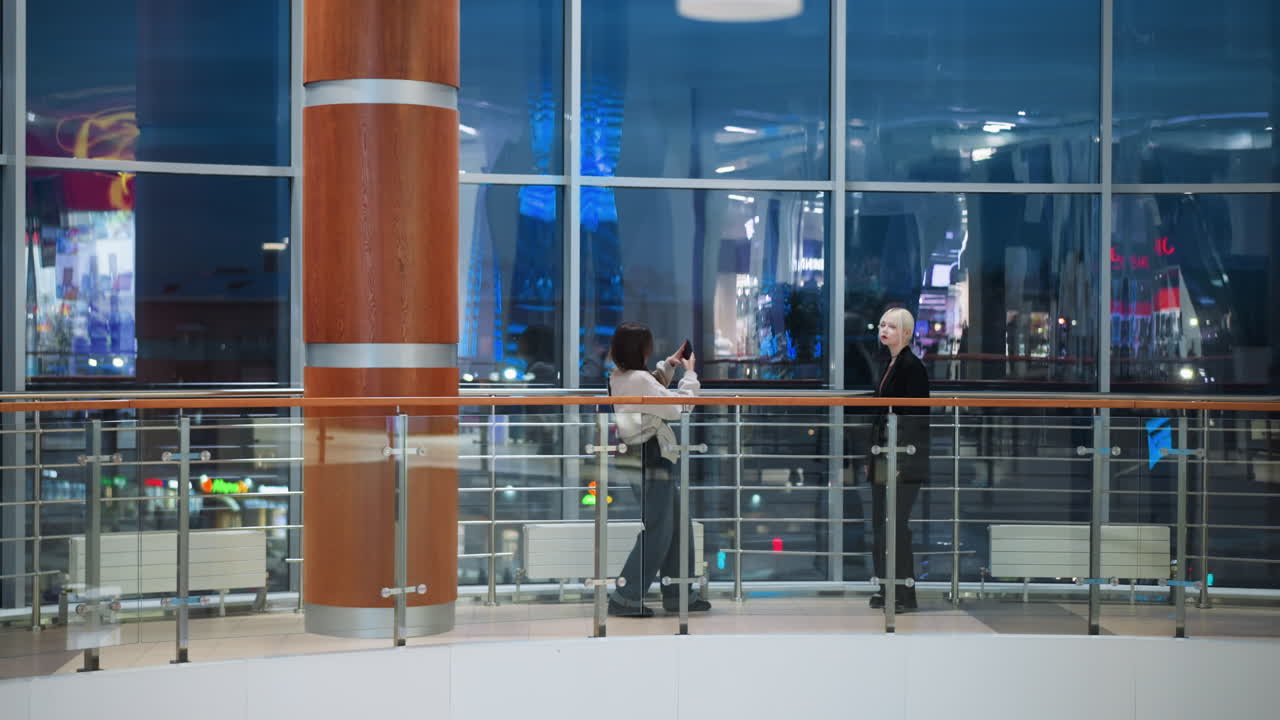 Two friends indoors in modern building near glass railing, one holding phone capturing photo of other against large glass windows with city lights reflecting