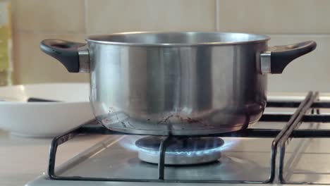 Stainless-steel-pot-heating-on-fire-ring-on-gas-stove---food-preparation-in-kitchen