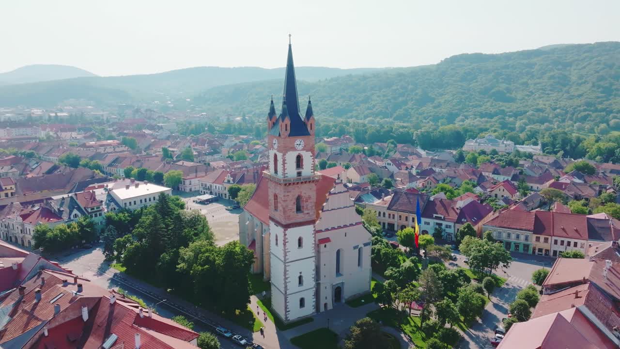 A aerial drone shot performs a slow orbit around the iconic Bistrita Evangelical Church tower, revealing the surrounding historic European city center and distant forested mountains