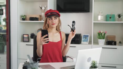 a young woman with light hair sitting at a table in a red dress,holding a phone in front of her,taking off her glasses,and mysteriously contemplating while looking in different directions