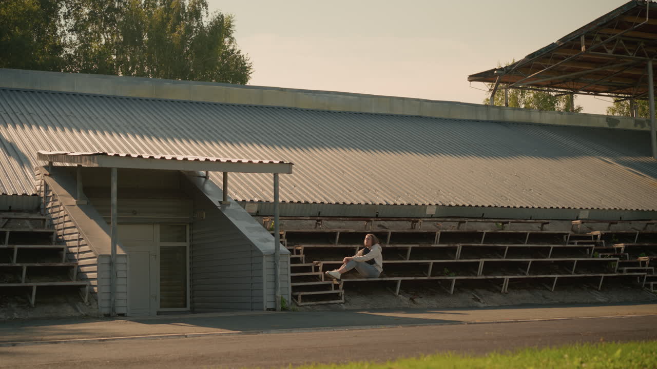 dama sentada sola en las gradas del estadio en una postura reflexiva con las manos en la pierna, mirando a la distancia con el techo de metal del estadio y el fondo natural con árboles y vegetación visibles
