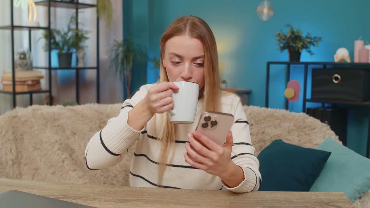 Caucasian woman scrolling social media feed on smartphone at home sipping coffee tea looking calm