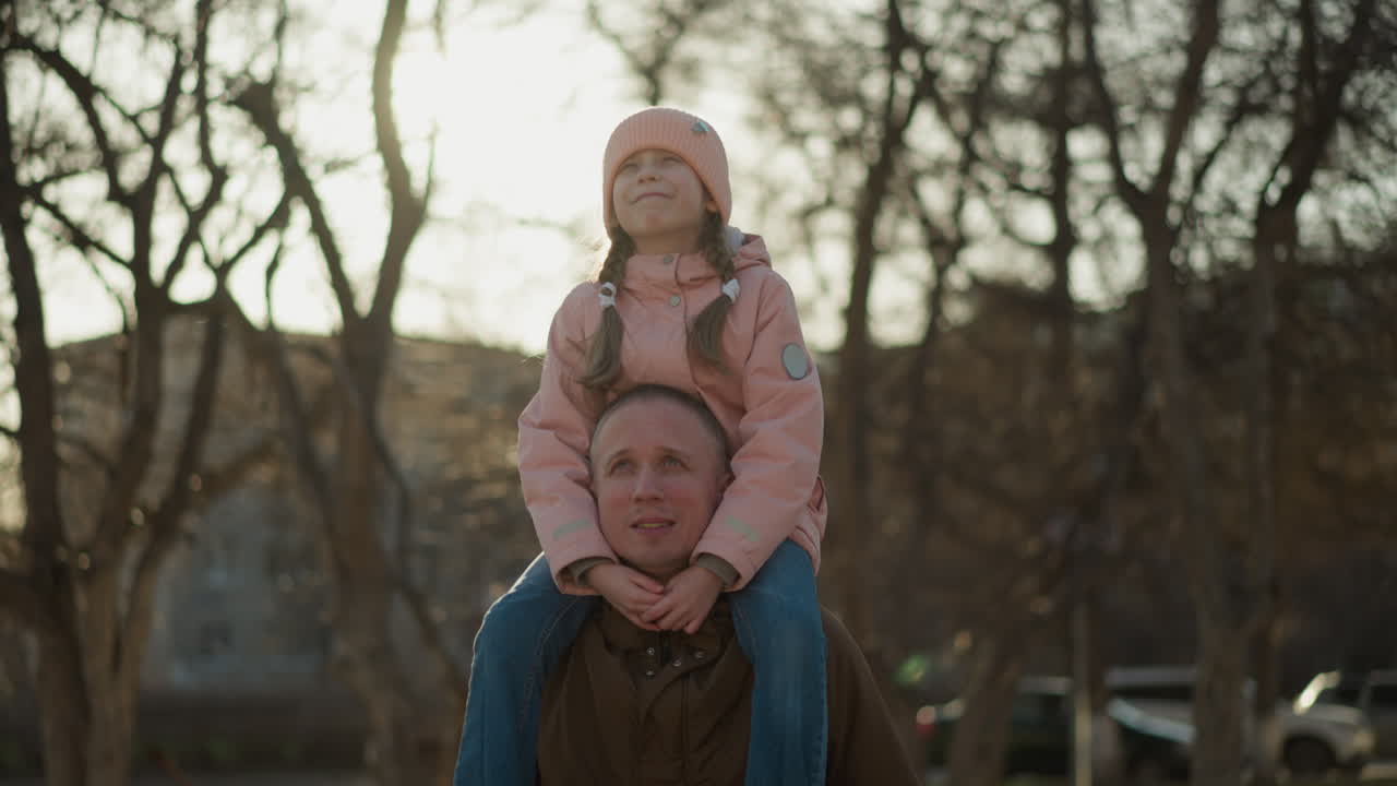 Front view of a little girl in a pink cap and jacket sitting joyfully on her dad's neck as they walk through a sunlit park. The girl is smiling with her eyes open, enjoying the moment