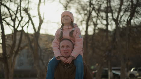 front view of a little girl in a pink cap and jacket sitting joyfully on her dad's neck as they walk through a sunlit park. the girl is smiling with her eyes open, enjoying the moment