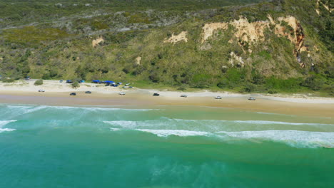 Drone-Aéreo-De-4k-De-Convoy-De-Automóviles-Conduciendo-En-Una-Playa-De-Arena-Junto-A-Las-Olas-Del-Océano-Azul