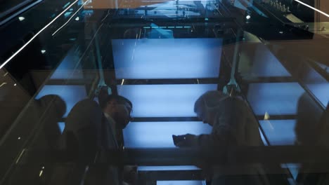 cinematic shot of modern glass elevator with group of businessmen move up. successful people take the lift to the top of business center. stylish man and woman talking while using smartphone.