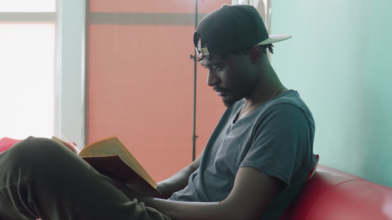 Seitenansicht eines jungen Mannes in Freizeitkleidung, der auf einem roten Sofa sitzt und mit konzentriertem Gesichtsausdruck ein Buch liest, eine verkehrt herum sitzende Mütze trägt, in einem hell erleuchteten Raum mit weichem, natürlichem Licht und pastellfarbenen Wänden