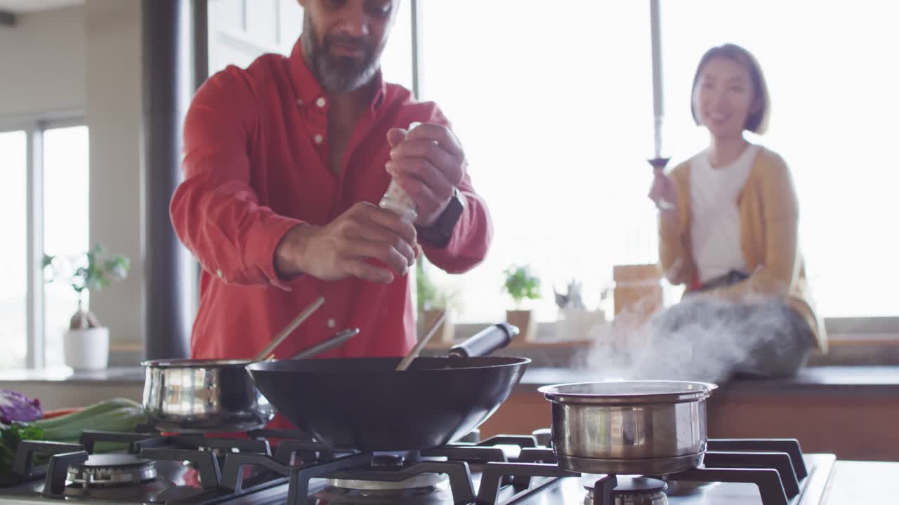 una pareja feliz y diversa cocinando juntos, sazonando comida en sartén en la cocina