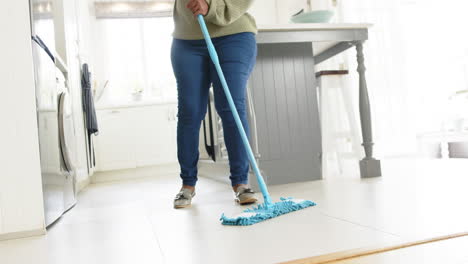 Feliz-Mujer-Mayor-Afroamericana-Limpiando-El-Piso-Y-Sonriendo-En-Una-Cocina-Soleada,-Cámara-Lenta