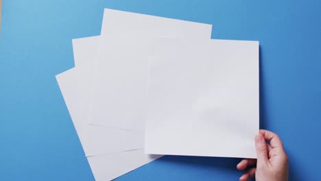 Hand-holding-piece-of-paper-over-pieces-of-paper-with-copy-space-on-blue-background-in-slow-motion