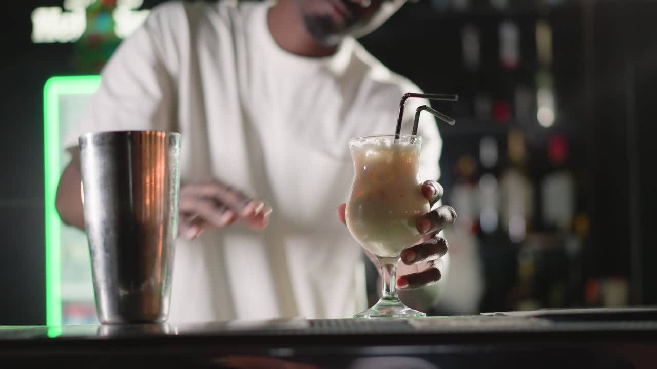 Bartender placing two straws into cocktail glass, preparing drink to serve. Behind, illuminated Heineken neon sign and well-stocked bar shelves, enhancing vibrant bar environment