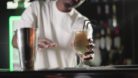 bartender placing two straws into cocktail glass, preparing drink to serve. behind, illuminated heineken neon sign and well-stocked bar shelves, enhancing vibrant bar environment