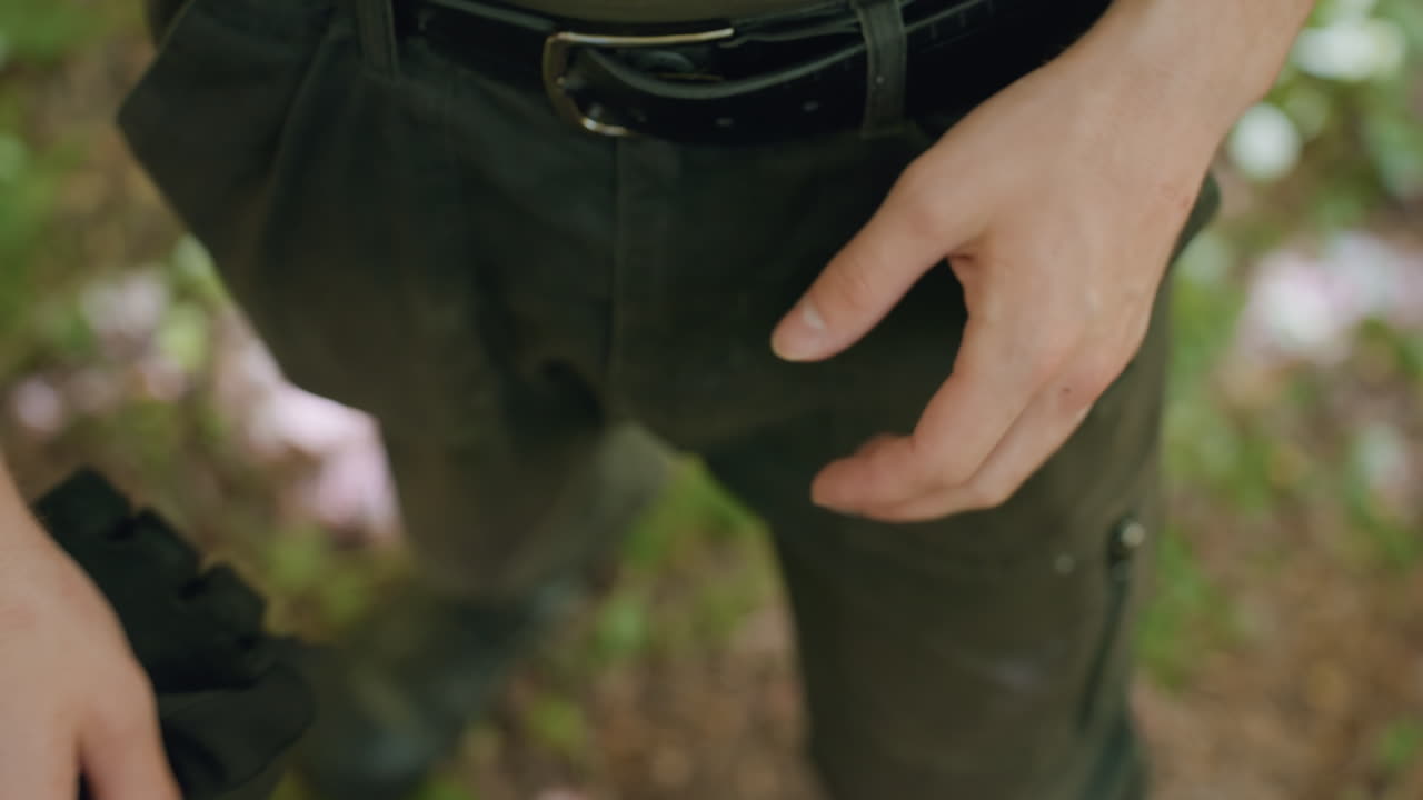 Close up view of tourist pulling off black fingerless glove at waist, belt buckle and cargo pants visible, veins on forearm highlighted by dappled sunlight through forest foliage, motion captured