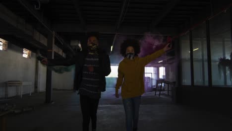 Mixed-race-man-and-woman-holding-blue-and-purple-flares-running-through-an-empty-building