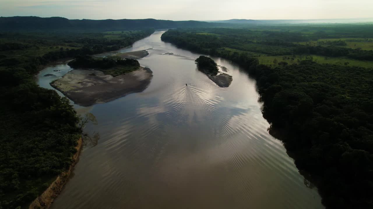 un bote a motor con pasajeros navega lentamente por el río hasta el horizonte desde las colinas colombianas al atardecer - sigue el disparo de un dron