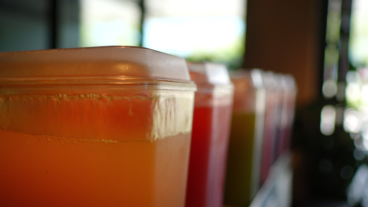 Orange juice and various colorful beverages filling plastic containers, arranged in dispensers, showcasing vibrant refreshment options at food service station with multiple drink choices